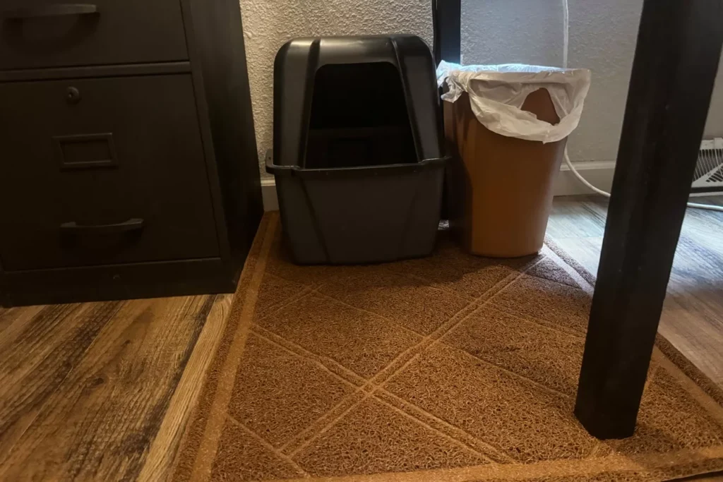 The 7 Best Cat Litter Mats for a Cleaner Home (Tested by Real Cats) 16 A brown, textured litter mat with a geometric diamond pattern placed on a wood-grain floor in front of a grey hooded cat litter box. A brown trash can with a white liner is to the right of the litter box.