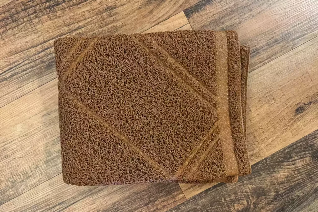 The 7 Best Cat Litter Mats for a Cleaner Home (Tested by Real Cats) 13 Overhead view of a brown, textured, spaghetti-loop style mat, folded into a square, resting on a dark wood-grain floor. The mat features a subtle geometric diamond pattern.
