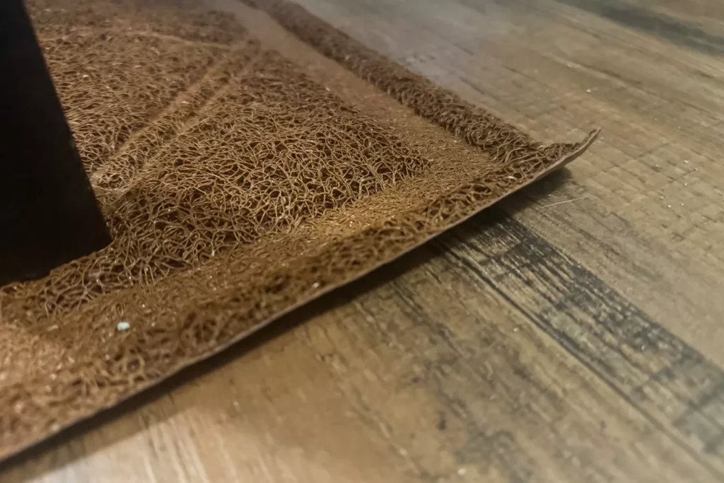 The 7 Best Cat Litter Mats for a Cleaner Home (Tested by Real Cats) 14 Close-up, low-angle shot of the edge of a brown textured mat on a wood-grain floor, highlighting its looped vinyl construction and a few tiny white specks, likely litter.
